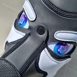 May include: Close-up of a black and white shoe with a unique design. The shoe features a black upper with white accents and a textured toe area. The design includes a colorful eye graphic with blue, pink, and purple hues.