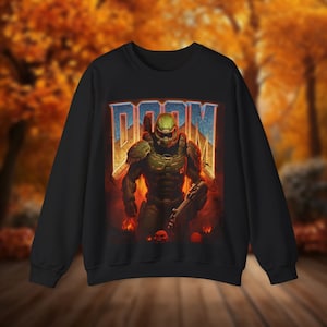 May include: Black sweatshirt featuring a graphic print of a green and black video game character holding a gun. The character is standing in front of a fiery background with the word "DOOM" in large letters above the character.