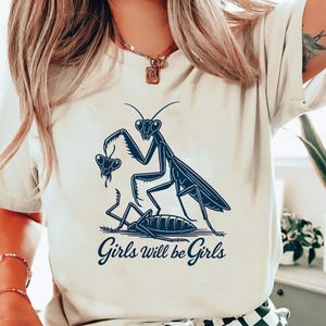 May include: Cream-colored t-shirt featuring a navy blue graphic of a praying mantis holding a smaller mantis, with a third mantis on the ground. The text "Girls will be Girls" is below the illustration. The shirt has short sleeves.