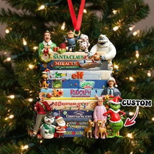 Puede incluir: Adorno navideño con títulos de películas como "The Santa Clause", "Elf" y "Home Alone". El adorno incluye figuras de personajes y una cinta roja para colgar. La palabra "CUSTOM" también es visible.