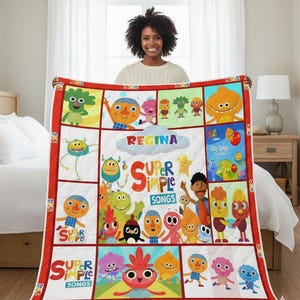 May include: A vibrant quilt displaying cartoon characters and the text "Super Simple Songs" and "Regina." The quilt has a red border and a white background with various square panels. The quilt is being held up by a person.