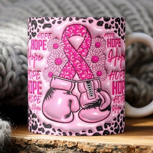 以下が含まれることがあります： ピンクのリボン、ボクシンググローブ、デイジーが描かれた白いセラミックマグ。デザインの周りには「HOPE」の文字が繰り返されています。背景は白黒のヒョウ柄です。