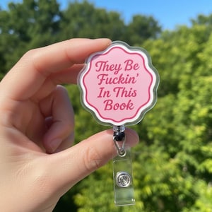 May include: A pink and white badge reel with the text "They Be Fuckin' In This Book". The badge reel has a clear plastic strap and a black bead.