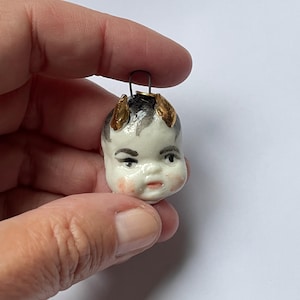 Peut inclure: Une petite tête de poupée ancienne en porcelaine avec des cheveux noirs et blancs et des cornes dorées. La tête de poupée a un visage peint avec des joues roses et une bouche rouge. Une boucle métallique est attachée en haut.