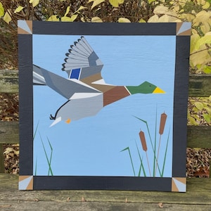 May include: A geometric painting of a flying duck against a light blue backdrop. The duck features shades of grey, brown, green, and yellow. The artwork includes reeds and grass, framed by a black border with corner accents.