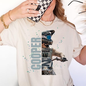 May include: A beige t-shirt with a graphic print of a motocross rider wearing a helmet and racing gear. The rider's name, "Cooper Webb", is printed vertically on the left side of the graphic. The graphic is surrounded by a light blue and green splatter pattern.