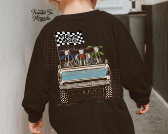 Può includere: Maglietta nera a maniche lunghe con un disegno di un cassone di camion azzurro chiaro che trasporta tre moto blu, rosse e verdi. La maglietta ha il testo "OFF TO THE RACES!" su una bandiera a scacchi. Per bambini.