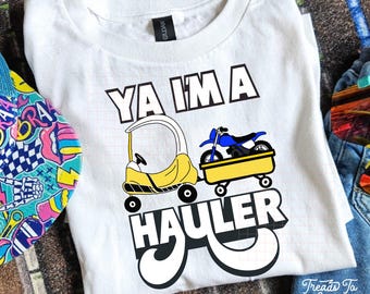 May include: White t-shirt with a graphic that says "YA I'M A HAULER". The graphic features a yellow toy car pulling a yellow trailer with a blue motorcycle. The shirt is made of a soft material.