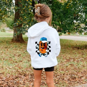 May include: White hoodie with a cartoon graphic on the back. The graphic features an orange character wearing a blue hat, set against a black and white checkered background. The hoodie has a hood and is worn by a person.