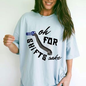 May include: Light blue t-shirt with the phrase "oh FOR SHIFTS sake" and a graphic of a black gear shift lever with blue accents. The shirt is a casual, oversized fit.
