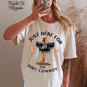 May include: Cream-colored t-shirt with the text "JUST HERE FOR THE DIRT COWBOY" and a graphic of a woman in a cowboy hat and fringe jacket. The shirt also features lightning bolt graphics. The brand name "Treads To Threads" is in the upper left corner.