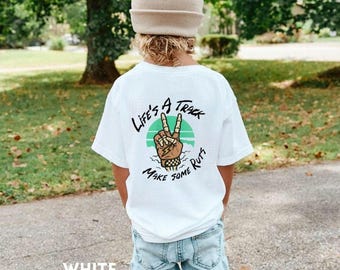 Può includere: T-shirt bianca con grafica sul retro raffigurante una mano che fa il segno della pace con un fulmine, un cerchio verde e la scritta "Life's a Track Make Some Ruts". La maglietta è indossata da un bambino.