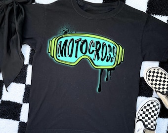 Può includere: T-shirt nero con grafica verde neon e blu di occhiali da motocross e la scritta "MOTOCROSS". La maglietta è esposta su una superficie a scacchi bianchi e neri, con un fiocco nero e scarpe slip-on a scacchi.