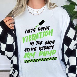 May include: White sweatshirt with the text "Cuts Down Vibration In The Bars Helps Reduce Arm Pump" in black and green. The design includes a nail polish bottle and a checkered pattern.