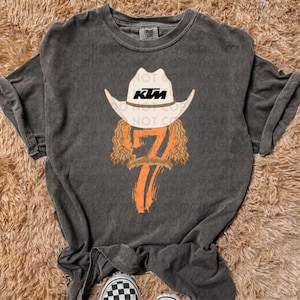 May include: A gray t-shirt with a graphic of a cowboy wearing a white cowboy hat with the word "KTM" on it. The cowboy has a mustache and long hair. The number "7" is printed in orange below the cowboy.