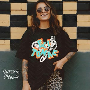 May include: Black t-shirt with the text "ELI TOMAC" in a colorful, 3D design, featuring a motorcycle graphic. The shirt is paired with leopard print pants. The brand "Treads Ta Threads" is printed on the bottom left.