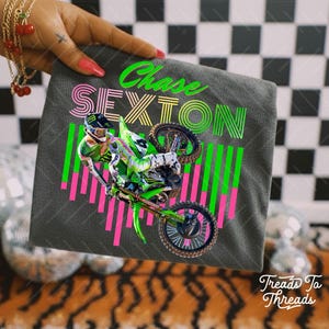 May include: A gray zippered pouch featuring a motocross rider in action. The design includes the name "Chase Sexton" in neon green and pink, with a matching graphic of vertical lines. The pouch is held up by a hand.
