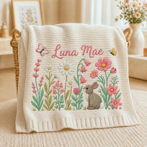 May include: A cream-coloured knit blanket featuring an embroidered design of a grey rabbit, various pink and white wildflowers, and two butterflies. The name Luna Mae is embroidered in pink script text above the floral and rabbit scene.