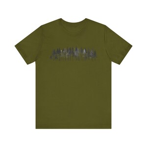 Könnte beinhalten: Ein olivgrünes Kurzarm-T-Shirt mit einem mittigen Grafikdruck einer dichten Waldlinie. Die Illustration zeigt verschiedene Kiefern in den Farben Dunkelgrau, Schwarz und Weiß, die eine geschichtete Waldsilhouette auf der Brust erzeugen.
