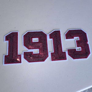 Op de afbeelding: Bordeauxrood pailletten nummerpatch met het jaartal "1913" in een collegiale lettertype. De cijfers zijn wit omlijnd. De patch staat op een lichtbeige achtergrond. Waarschijnlijk voor een jas of kleding.