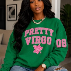 Puede incluir: Una sudadera verde de cuello redondo con el texto PRETTY VIRGO en letras rosas de estilo universitario en el pecho. Debajo del texto hay un gráfico de una hoja rosa, y el número 08 aparece en rosa en la manga izquierda. Mangas largas.