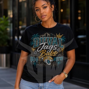 May include: A black short-sleeve t-shirt featuring a graphic print with the text "DUVAL Jags Babe", "904", and "#DTWD". The design includes a jaguar head, palm trees, a crown, and a leopard print background in shades of teal, gold, and black.