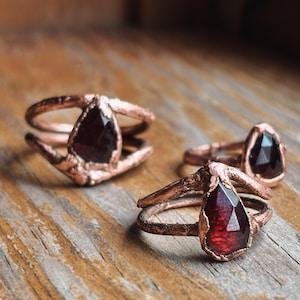 May include: Close-up of three copper rings with teardrop-shaped, faceted, dark red gemstones. The rings have a rustic, textured appearance, set on a wooden surface. The gemstones are the focal point, catching the light and adding a touch of elegance.