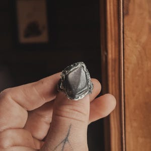 Könnte beinhalten: Ein silberner Ring mit einem großen, unregelmäßig geformten, grauen Edelstein. Der Ring hat ein strukturiertes, organisches Design um den Stein herum. Der Ring wird von einer Hand mit einem kleinen Tattoo am Finger gehalten.