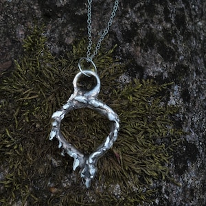 May include: A silver-colored pendant necklace with a unique, organic design. The pendant is a ring-like shape with textured, irregular edges, and is suspended from a delicate chain. The necklace is displayed on a mossy, dark background.