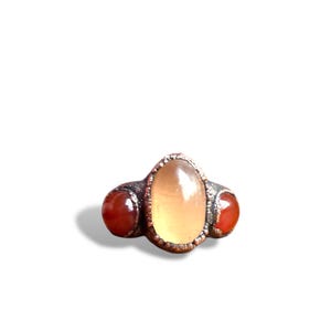 May include: A silver-toned ring featuring three gemstones. The central stone is oval-shaped and light yellow, flanked by two round, reddish-orange stones. The ring has a textured band and a vintage aesthetic.