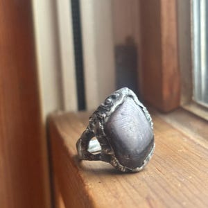 Puede incluir: Un anillo de plata con una gran piedra preciosa ovalada gris. La banda y el engaste del anillo tienen un diseño texturizado y ornamentado. El anillo se exhibe sobre una superficie de madera, con una ventana y molduras de madera en el fondo.