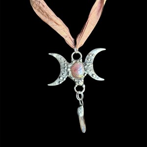 May include: A silver-toned pendant necklace featuring a triple moon design with a central oval-shaped stone and a dangling crystal. The pendant is suspended from a light brown ribbon. The background is black.