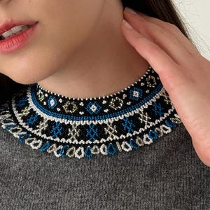 May include: A beaded choker necklace with a geometric design. The necklace features black, white, blue, and gold beads. The necklace is worn over a grey jumper. A silver hoop earring is visible.