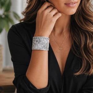 May include: A wide cuff bracelet featuring a textured gray and white animal hide pattern with a smooth metallic silver border. The bracelet has a rigid, curved structure that wraps around the wrist, showcasing a distinct mottled fur-like appearance.