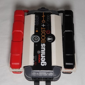May include: A white and black portable car jump starter with the text "genius" and "BOOST +" printed on the front. The jump starter has a red and black cable attached to it. The jump starter has a USB port and a 12V 1000A output.