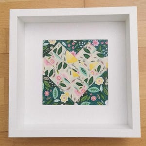 Puede incluir: Un marco cuadrado blanco que contiene una obra de arte con forma de corazón formada por tiras de tela. El corazón presenta un patrón de limones amarillos, hojas verdes y pequeñas flores rosas y amarillas sobre un fondo blanco, sobre un fondo floral verde oscuro.