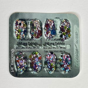 May include: A blister pack of Paracetamol 500 mg tablets, decorated with colorful beads and glitter. The text "Paracetamol 500 mg Tablets" is visible. The expiration date is June 2027.