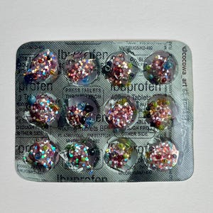 May include: A silver blister pack of ibuprofen tablets, each cavity filled with colorful glitter and beads. The word "Ibuprofen" is visible on the packaging, along with other text. The artwork is set against a white background.