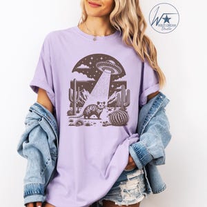 K&ouml;nnte beinhalten: Ein lavendelfarbenes T-Shirt mit einem Aufdruck eines Opossums, das in einer W&uuml;stenlandschaft mit Kakteen steht, &uuml;ber dem ein UFO schwebt. Das UFO sendet einen Lichtstrahl aus.