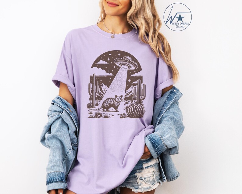 K&ouml;nnte beinhalten: Ein lavendelfarbenes T-Shirt mit einem Aufdruck eines Opossums, das in einer W&uuml;stenlandschaft mit Kakteen steht, &uuml;ber dem ein UFO schwebt. Das UFO sendet einen Lichtstrahl aus.