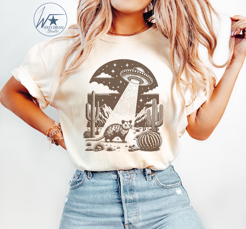 K&ouml;nnte beinhalten: Ein wei&szlig;es T-Shirt mit einem braunen Aufdruck eines Opossums in der W&uuml;ste mit einem UFO dar&uuml;ber. Der Aufdruck enth&auml;lt auch Kakteen und Berge.