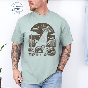 K&ouml;nnte beinhalten: Ein hellblaues T-Shirt mit einem grafischen Aufdruck eines Opossums, das in einer W&uuml;stenlandschaft mit Kakteen steht und ein UFO dar&uuml;ber schwebt. Das UFO sendet einen Lichtstrahl aus, der auf das Opossum scheint.