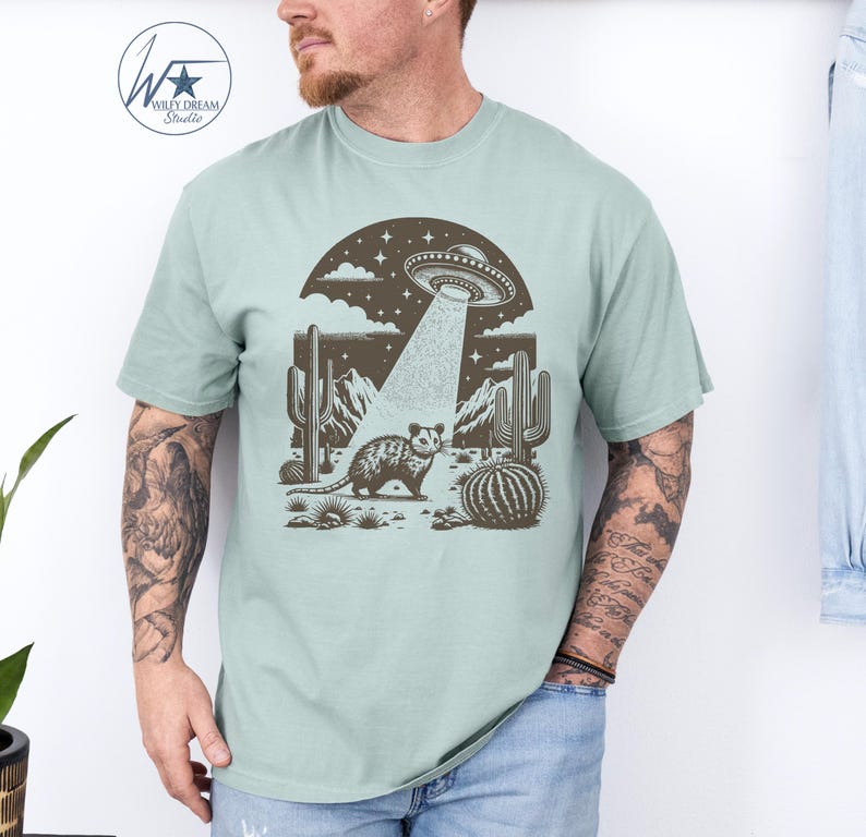 K&ouml;nnte beinhalten: Ein hellblaues T-Shirt mit einem grafischen Aufdruck eines Opossums, das in einer W&uuml;stenlandschaft mit Kakteen steht und ein UFO dar&uuml;ber schwebt. Das UFO sendet einen Lichtstrahl aus, der auf das Opossum scheint.
