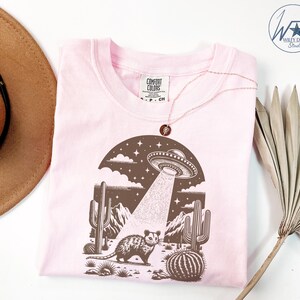 K&ouml;nnte beinhalten: Ein pinkfarbenes T-Shirt mit einem braunen Grafikdruck einer W&uuml;stenlandschaft mit einer fliegenden Untertasse, Kakteen und einem kleinen Tier. Der Text "COMFORT COLORS" befindet sich auf einem wei&szlig;en Etikett.