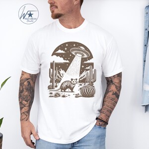 K&ouml;nnte beinhalten: Ein wei&szlig;es T-Shirt mit einer braunen Illustration eines Opossums in einer W&uuml;stenlandschaft mit Kakteen und einem UFO, das dar&uuml;ber schwebt. Das UFO sendet einen Lichtstrahl aus, der auf das Opossum scheint.