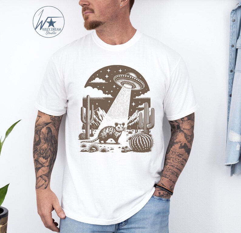 K&ouml;nnte beinhalten: Ein wei&szlig;es T-Shirt mit einer braunen Illustration eines Opossums in einer W&uuml;stenlandschaft mit Kakteen und einem UFO, das dar&uuml;ber schwebt. Das UFO sendet einen Lichtstrahl aus, der auf das Opossum scheint.