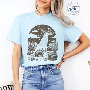 K&ouml;nnte beinhalten: Ein hellblaues T-Shirt mit einem Aufdruck eines Opossums, das in einer W&uuml;stenlandschaft mit Kakteen steht, &uuml;ber dem ein UFO schwebt. Das UFO sendet einen Lichtstrahl aus.