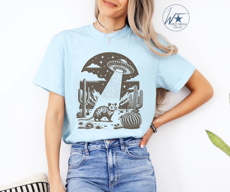K&ouml;nnte beinhalten: Ein hellblaues T-Shirt mit einem Aufdruck eines Opossums, das in einer W&uuml;stenlandschaft mit Kakteen steht, &uuml;ber dem ein UFO schwebt. Das UFO sendet einen Lichtstrahl aus.