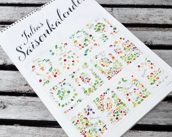 May include: A white calendar with the title "Julia's Saisonkalender" printed in black cursive script. The calendar features a monthly grid with each month represented by a colorful illustration of seasonal fruits and vegetables.