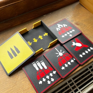 Pode incluir: Um conjunto de bases para copos quadradas com um esquema de cores preto, amarelo e vermelho, apresentando vários ícones gráficos, incluindo balas, fogo e setas. O conjunto inclui um suporte quadrado preto e amarelo a condizer.
